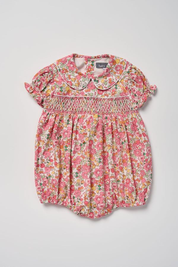 A toddler girl's short-sleeve floral romper in pink, red, and yellow with a smocked chest and a Peter Pan collar, shown flat on a light gray background.