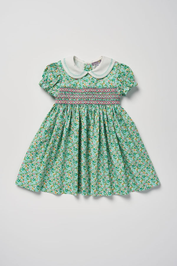 A toddler girl's short-sleeve dress with a green and white floral print, featuring a smocked bodice with pink and blue stripes and a white Peter Pan collar.